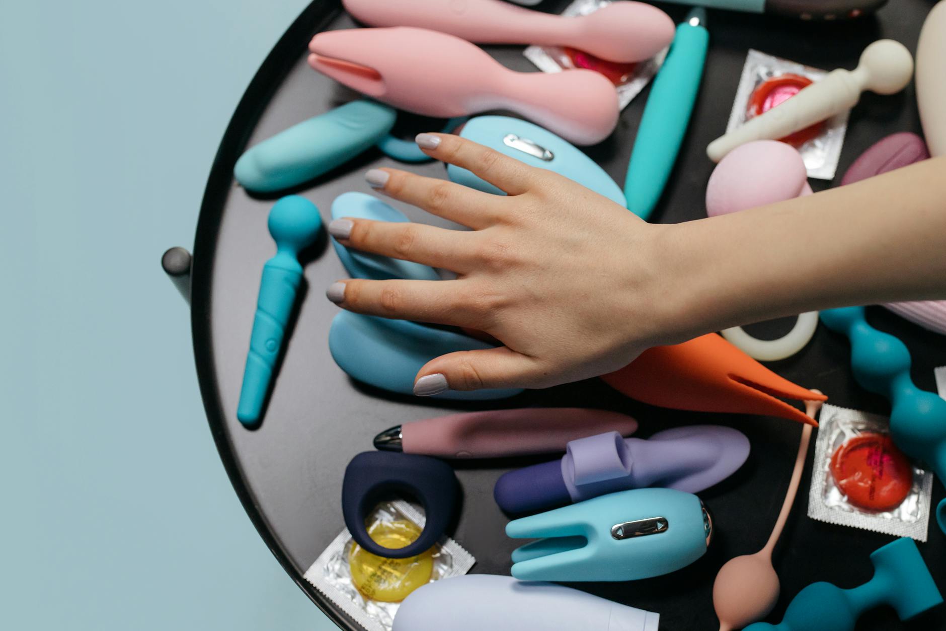 A hand reaching over a variety of colorful sex toys arranged on a table.