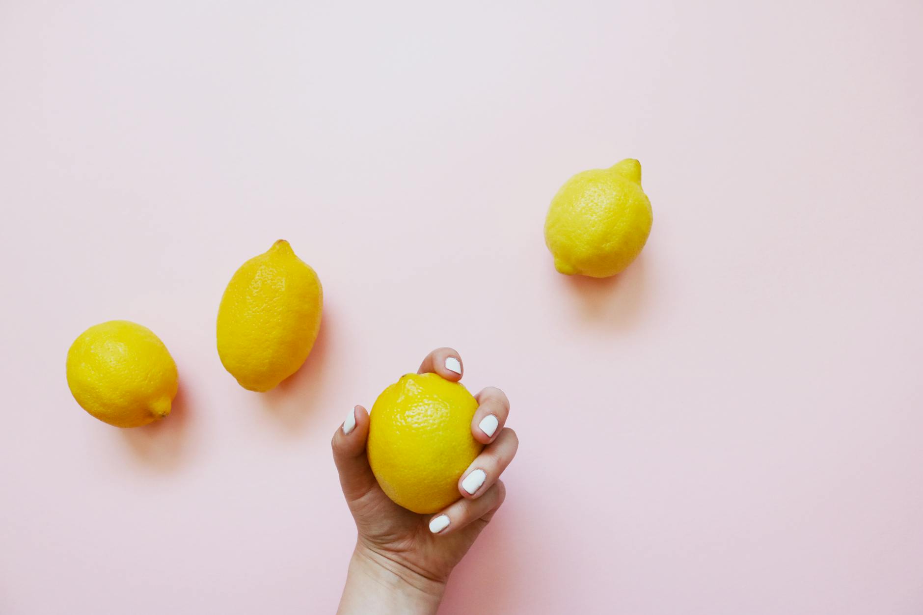 A hand with white nails holding a fresh lemon on a soft pink background with additional lemons nearby
