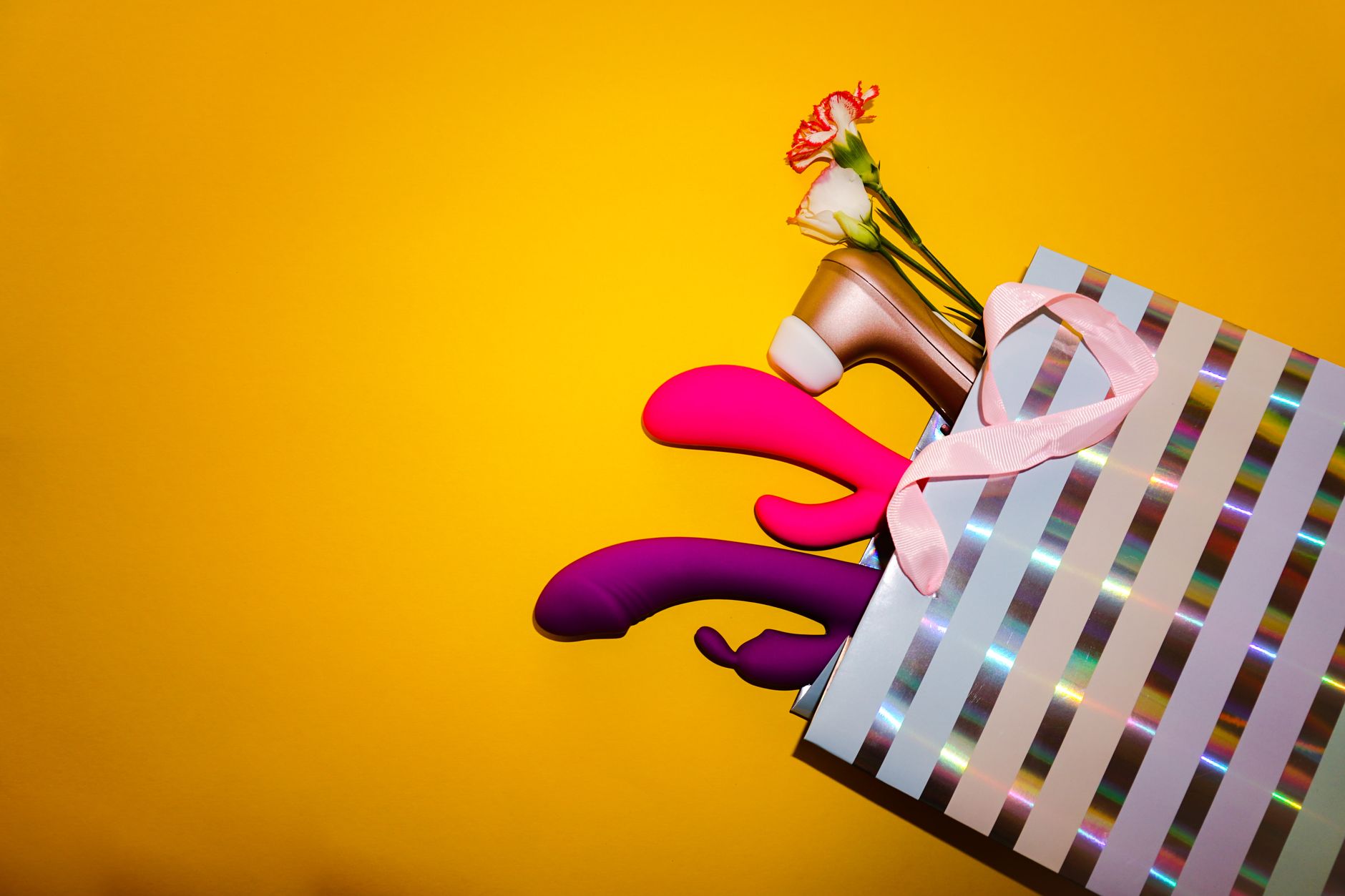 Colorful vibrators with flowers in a holographic gift bag, set against a bold yellow background.